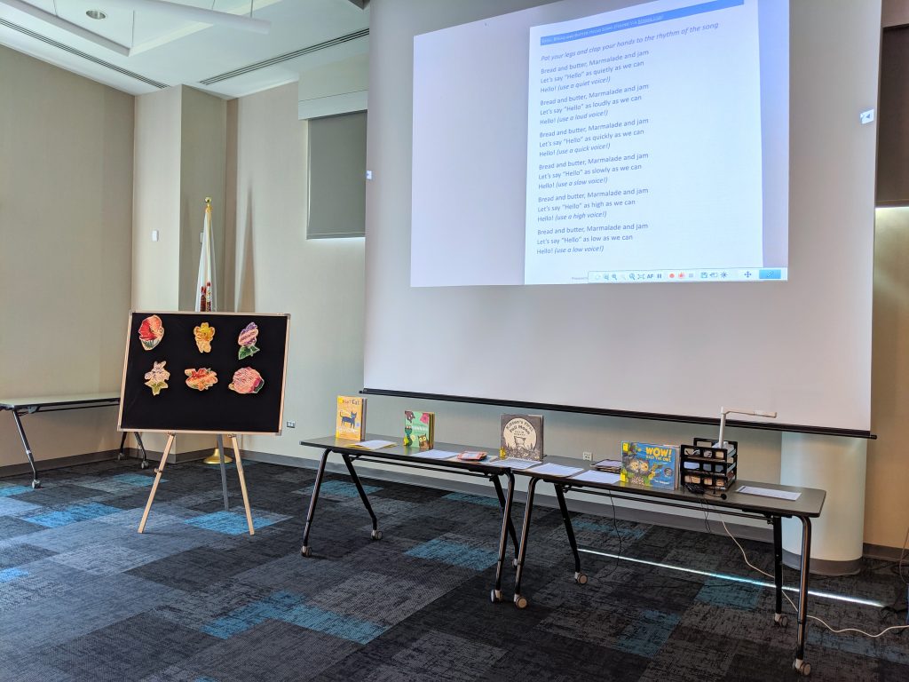 Naperville Public LIbrary Meeting Room set up for Family Storytime (projector screen pulled down, tables below screen with books and puppets, a flannel board off to the left for flannel stories)