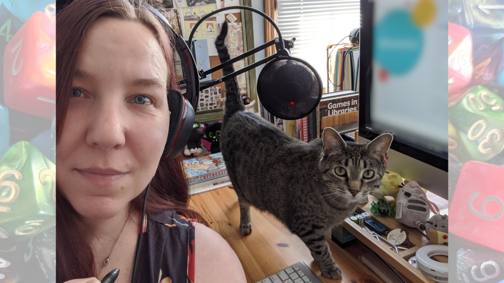 Selfie of Rebecca at her computer with her recording mic, there is a cat on the desk